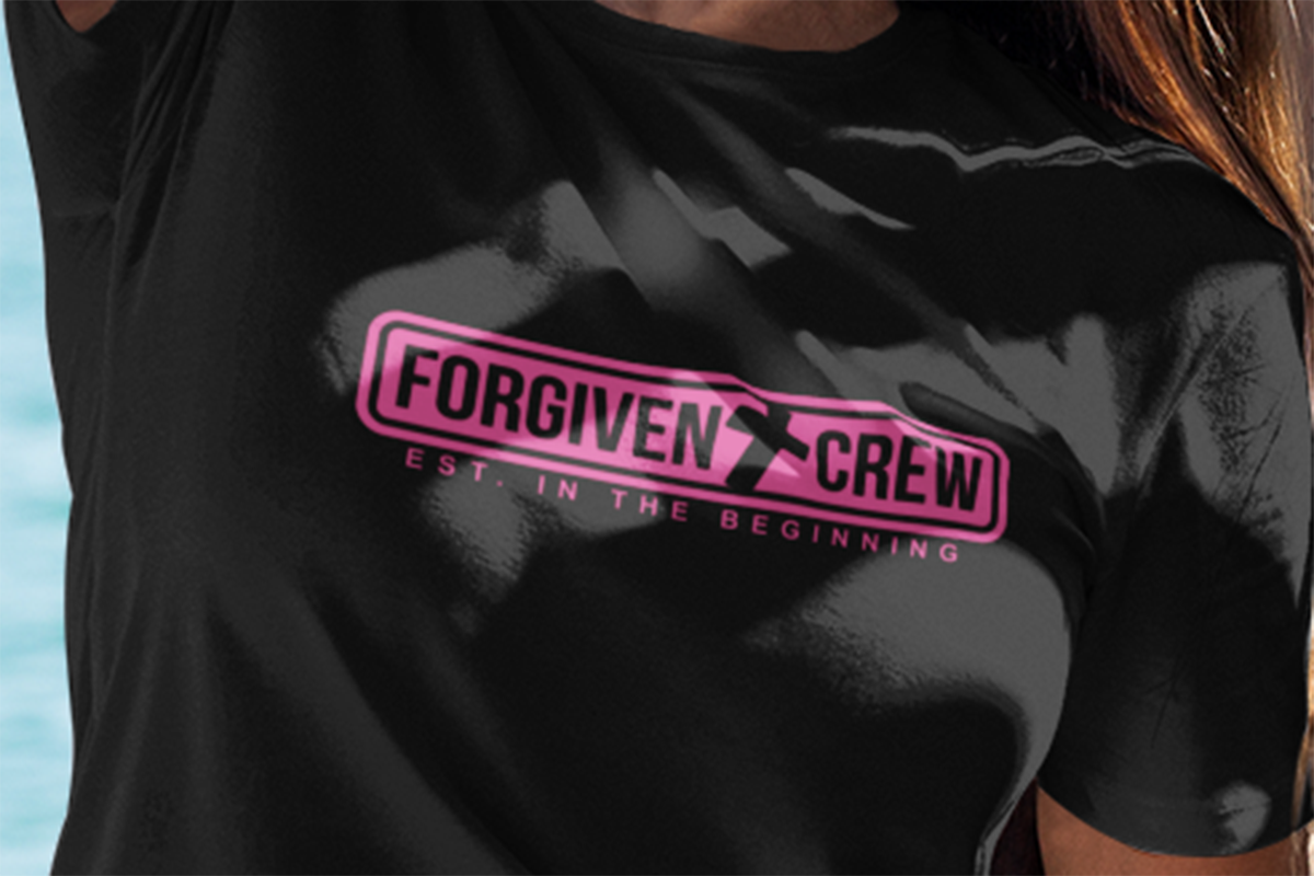 Close up version of the Picture of girl next to the ocean with sunglasses wearing a black Forgiven Crew Christian T-shirt printing in pink.